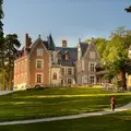 Une personne marchant sur un chemin menant à un grand manoir historique en briques rouges entouré de pelouses et d'arbres verts, par une journée ensoleillée.