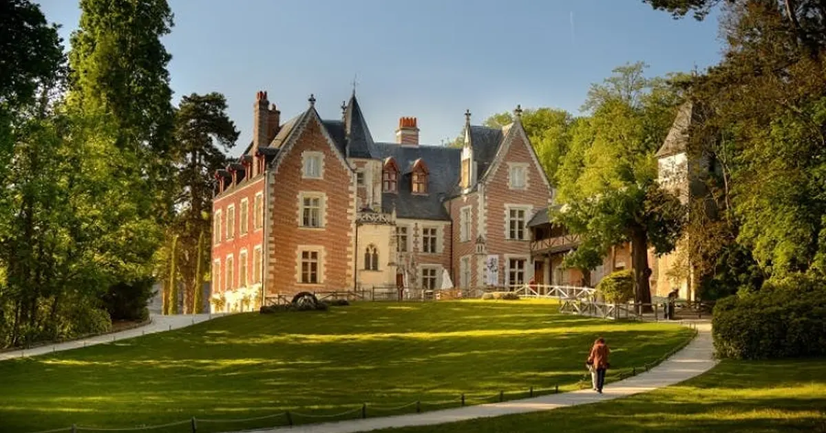 Une personne marchant sur un chemin menant à un grand manoir historique en briques rouges entouré de pelouses et d'arbres verts, par une journée ensoleillée.