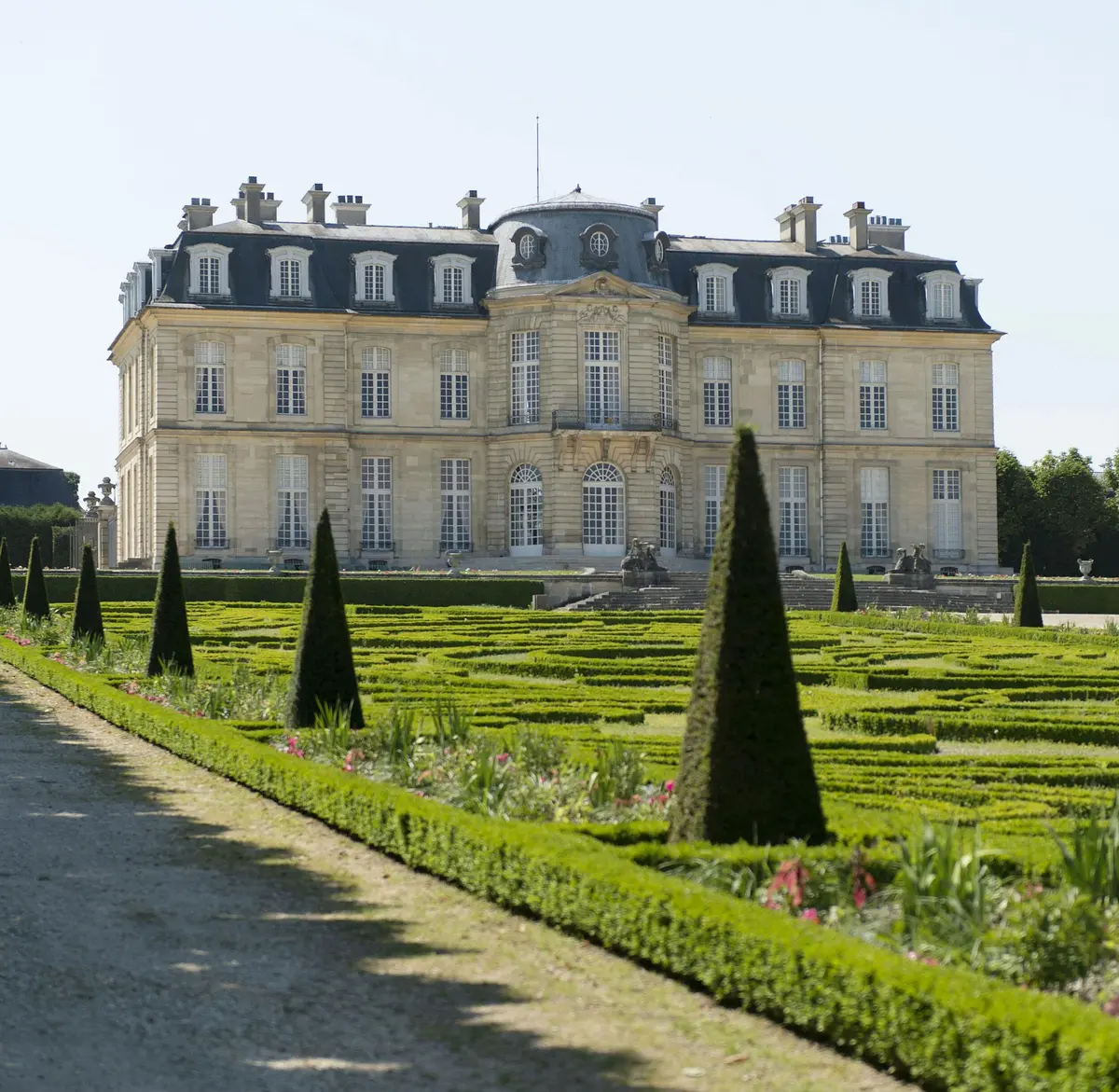 Jardin formel avec des haies soignées et des arbres topiaires devant une grande demeure ornée de multiples fenêtres et d'un toit pentu.