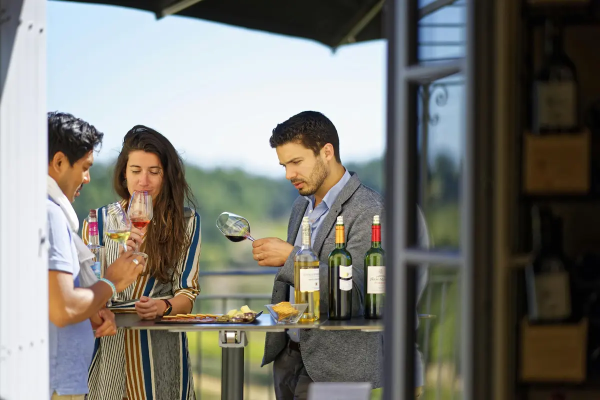 Trois personnes &agrave; une table en plein air d&eacute;gustant du vin, avec quatre bouteilles de vin et un plateau d'amuse-gueule. Arbres et ciel en arri&egrave;re-plan.