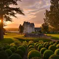 Jardin entretenu avec des haies rondes, des arbres et un bâtiment en pierre à l'arrière-plan au coucher du soleil.