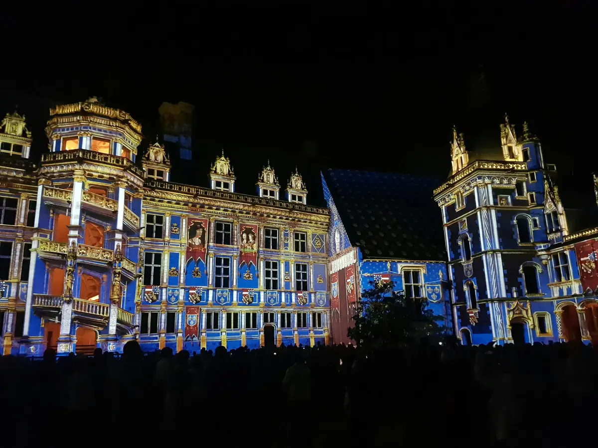 Un bâtiment illuminé par des lumières bleues, rouges et dorées dans un ciel nocturne sombre, avec une foule de personnes devant.