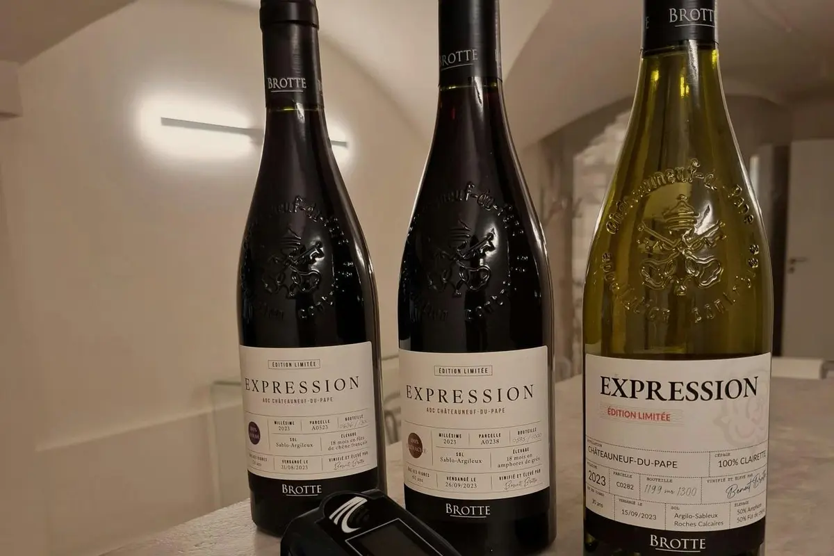 Trois bouteilles de vin Brotte étiquetées Expression, dont une jaune et deux foncées, placées sur une table à côté d'un appareil numérique.