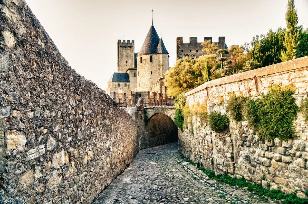 Un chemin pavé mène à un château historique en pierre avec plusieurs tours et un pont, flanqué de hauts murs en pierre.