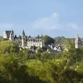 Vue panoramique d'un château historique et d'un village entouré d'arbres, avec un ciel bleu clair en arrière-plan.