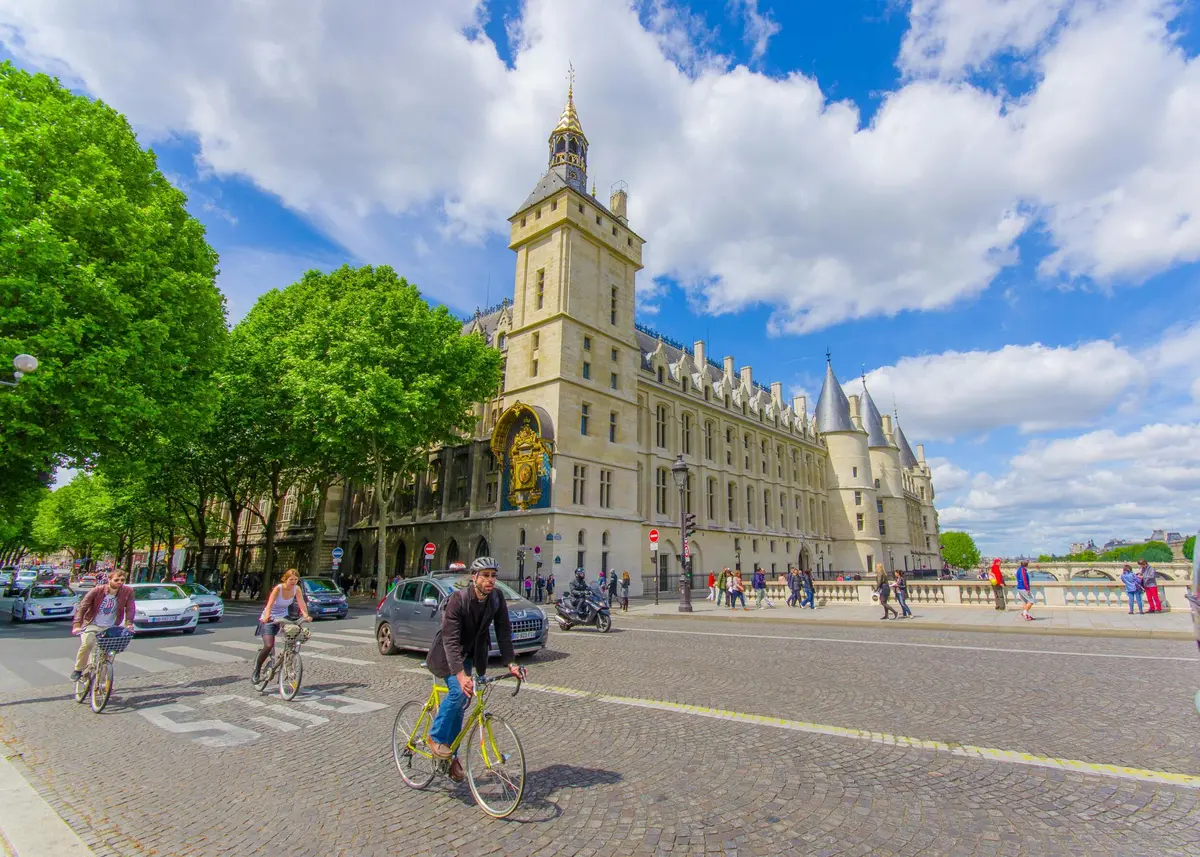 Des personnes à vélo et à pied près d'un bâtiment historique et orné d'une tour d'horloge et de tourelles pointues, sous un ciel lumineux et partiellement nuageux.