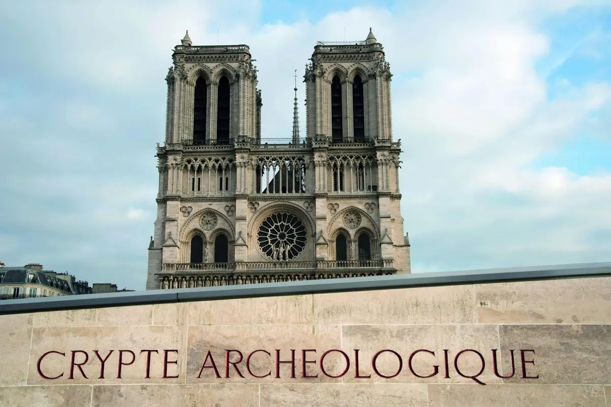 Façade de la cathédrale Notre-Dame avec ses tours jumelles et son vitrail, vue d'en dessous d'un panneau en pierre indiquant Crypte Archéologique.