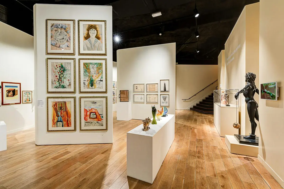 Galerie d'art comprenant des peintures encadrées sur les murs, des sculptures sur des piédestaux, un sol en bois et un escalier en arrière-plan.