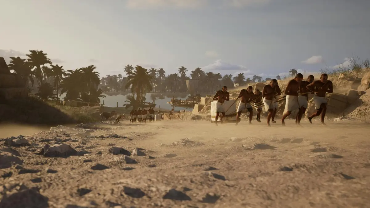 Des hommes en pagne tirent une lourde charge sur un chemin poussiéreux avec des palmiers et une rivière en arrière-plan.