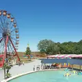 Grande roue colorée, bateaux tamponneurs dans une piscine et tente rayée entourée d'arbres sous un ciel bleu clair dans un parc d'attractions.