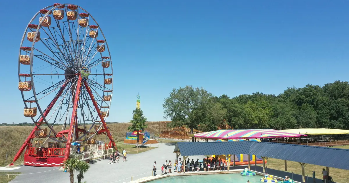 Grande roue colorée, bateaux tamponneurs dans une piscine et tente rayée entourée d'arbres sous un ciel bleu clair dans un parc d'attractions.