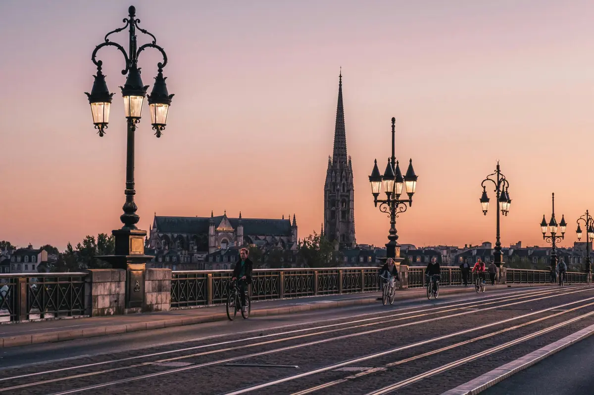 Des cyclistes roulent sur un pont orné de lampadaires au coucher du soleil, avec une haute flèche historique et une église en arrière-plan.