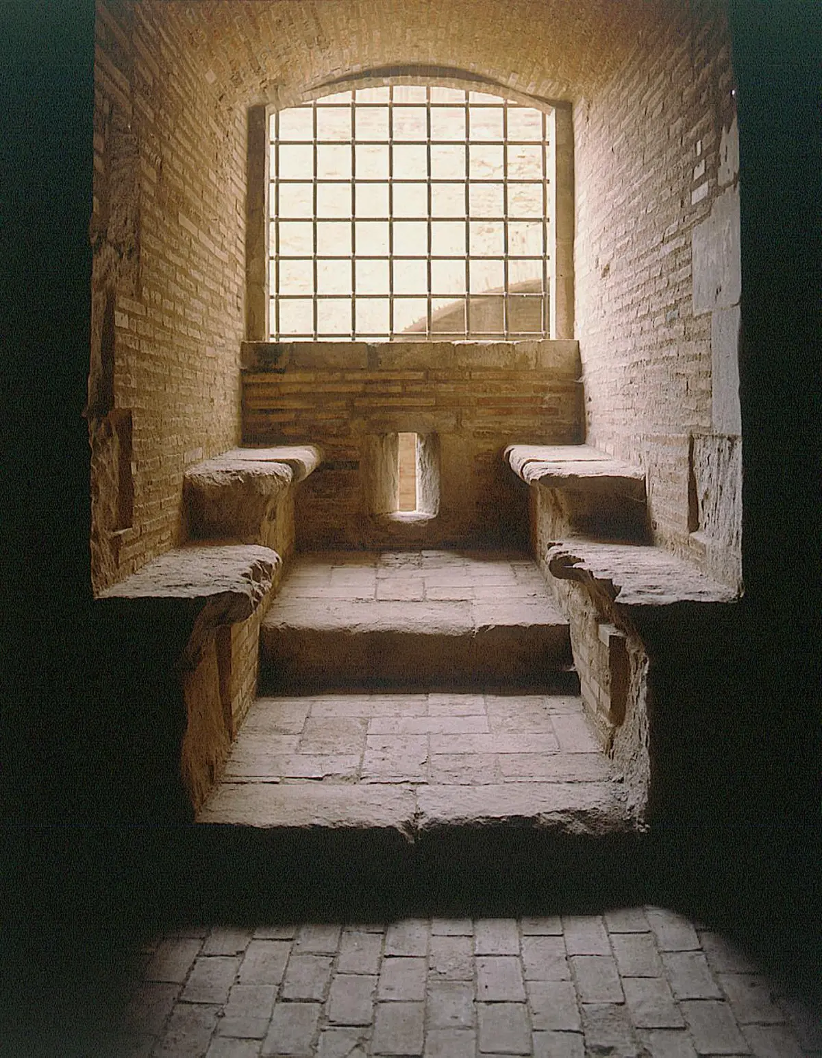 Une pièce étroite en pierre avec des marches arquées et deux bancs en pierre, éclairée par une petite fenêtre avec des barreaux en métal.