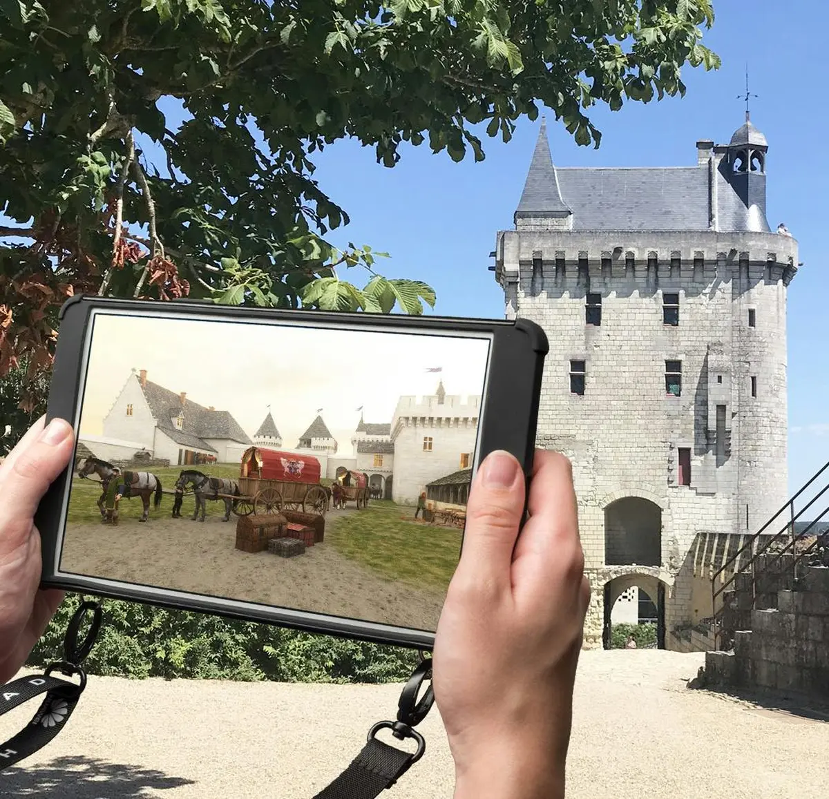 Une personne tient une tablette affichant une scène médiévale devant un véritable château médiéval sous un ciel bleu clair.