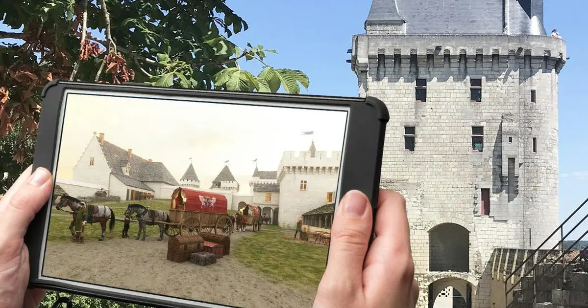 Une personne tient une tablette affichant une scène médiévale devant un véritable château médiéval sous un ciel bleu clair.