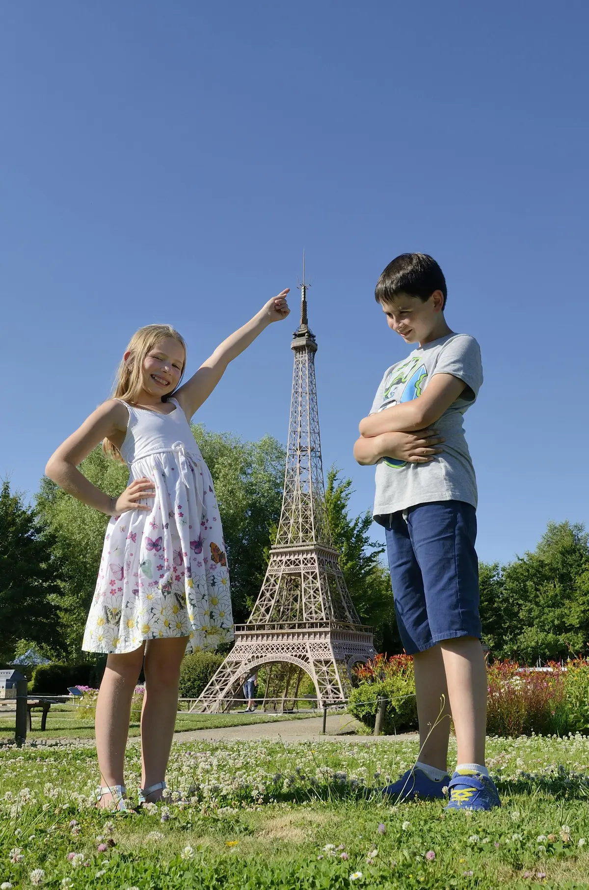 Une fille montre du doigt une tour Eiffel miniature en souriant, tandis qu'un garçon se tient à côté d'elle, les bras croisés, tous deux en plein air par une journée ensoleillée.