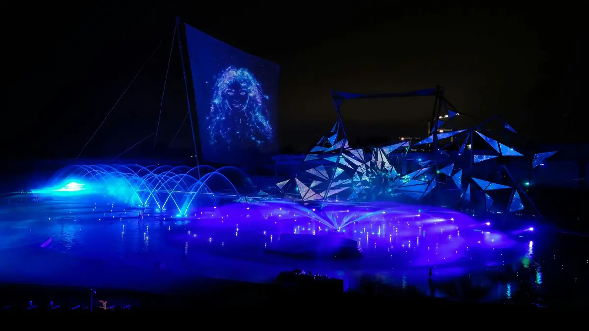 Un spectacle nocturne de lumière et d'eau avec des fontaines lumineuses, des structures abstraites et un visage projeté sur un grand écran.