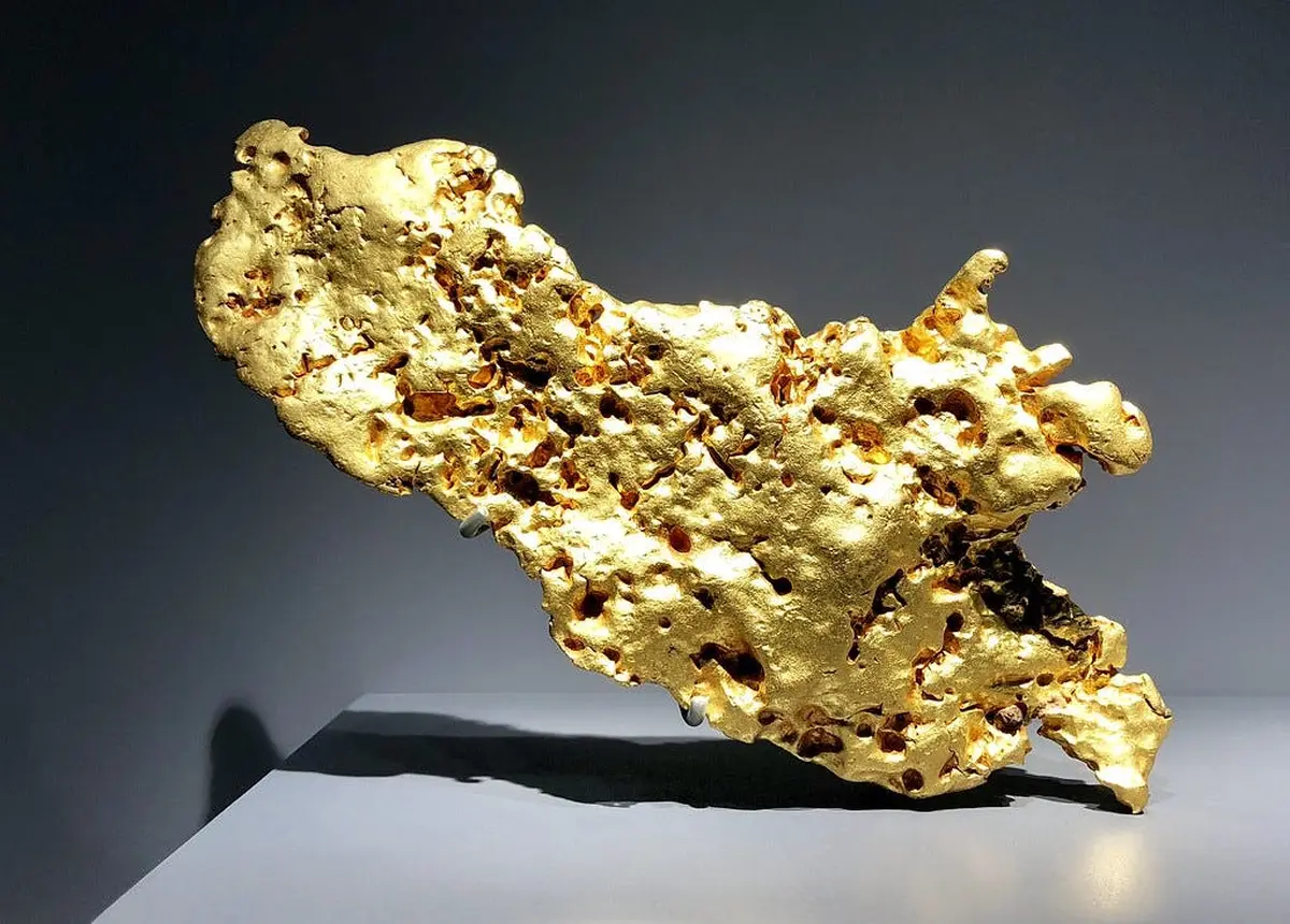 Grande pépite d'or de forme irrégulière présentée sur un socle gris sur fond sombre.