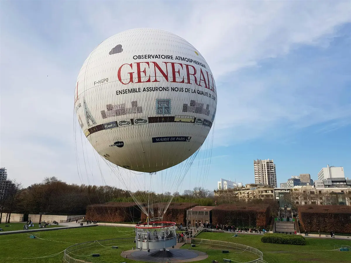 Grand ballon captif dans un parc, étiqueté GENERALI, avec des personnes rassemblées autour. Des bâtiments et des arbres sont visibles à l'arrière-plan.