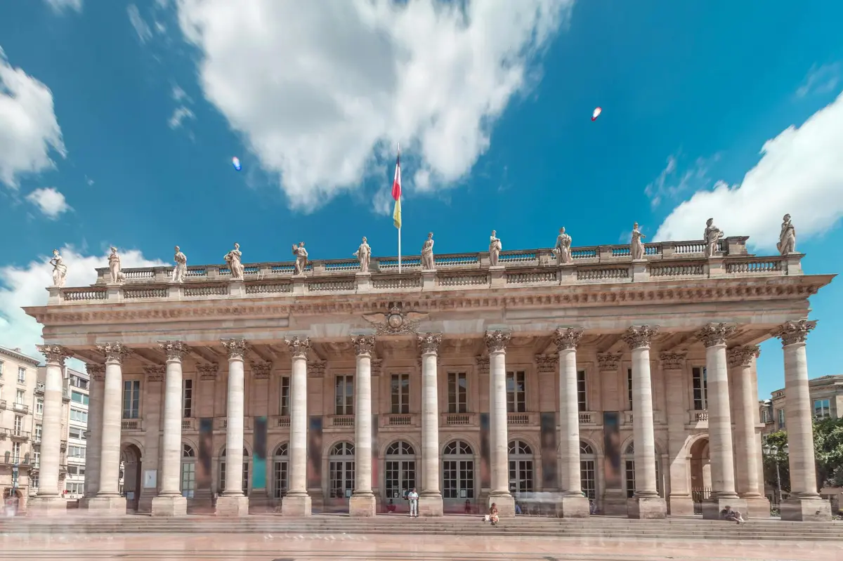 Un bâtiment néoclassique orné de hautes colonnes et de statues, surmonté d'un drapeau, sous un ciel bleu éclatant parsemé de nuages.
