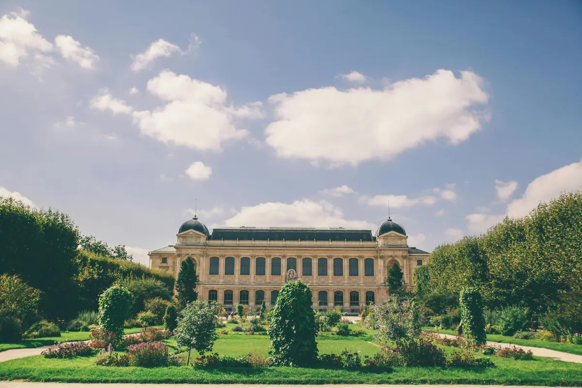 Grand bâtiment avec des tours en forme de dôme, entouré d'un jardin avec des fleurs et des arbustes, sous un ciel partiellement nuageux.