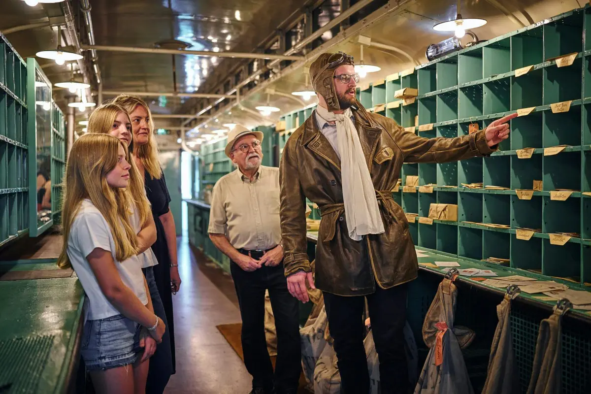Un groupe de personnes &agrave; l'int&eacute;rieur d'un wagon de train d'&eacute;poque : l'une d'entre elles, v&ecirc;tue d'un costume d'aviateur, montre quelque chose du doigt tandis que les autres l'observent attentivement.