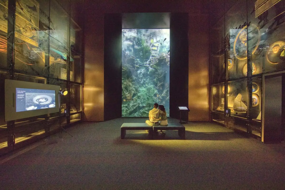 Un couple est assis sur un banc, face à un grand aquarium éclairé dans une galerie sombre avec des vitrines de chaque côté.
