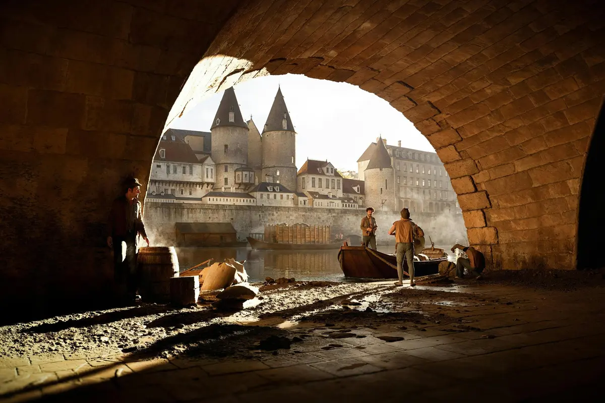Vue d'un front de mer sous une arche en pierre, avec des personnes près de bateaux et, en arrière-plan, une ville historique dotée de tours.