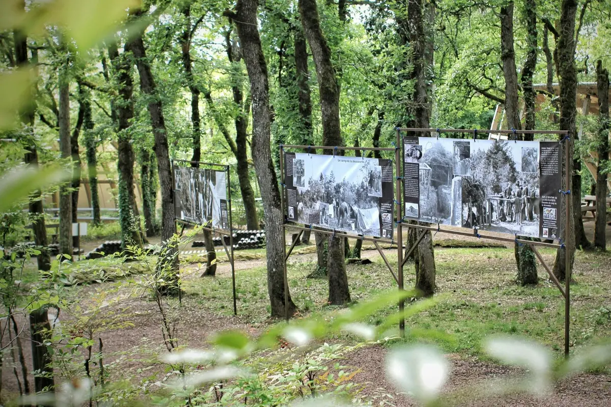 Exposition en plein air, dans une zone boisée, présentant de grands panneaux avec des photographies historiques en noir et blanc et des textes.