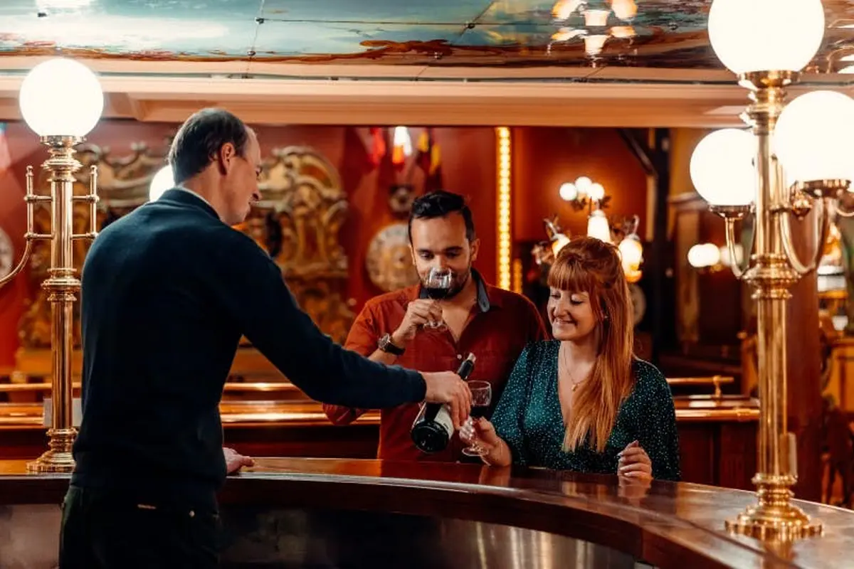 Trois personnes au bar : le barman verse du vin à un homme et à une femme. L'arrière-plan présente un éclairage chaleureux et un décor orné.
