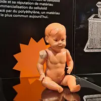 Une poupée en plastique usée et nue, dont l'épaule est abîmée, est présentée sur un fond noir avec un texte et des illustrations en français. &copy; Villa de Colmar