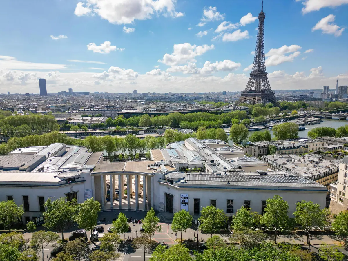 Vue aérienne de la Tour Eiffel et des bâtiments parisiens environnants avec de la verdure et un ciel partiellement nuageux.