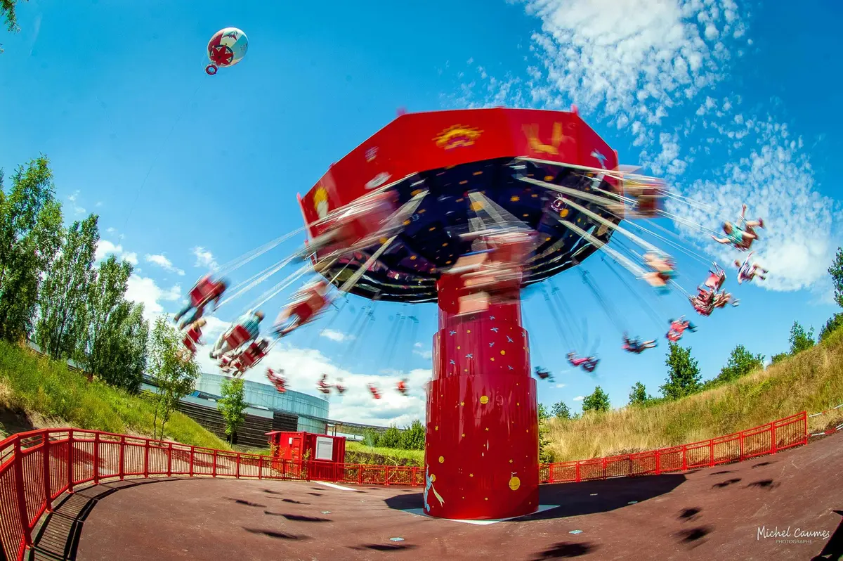 Un carrousel rouge en mouvement sur fond de ciel bleu avec des personnes assises et un ballon flottant à proximité.