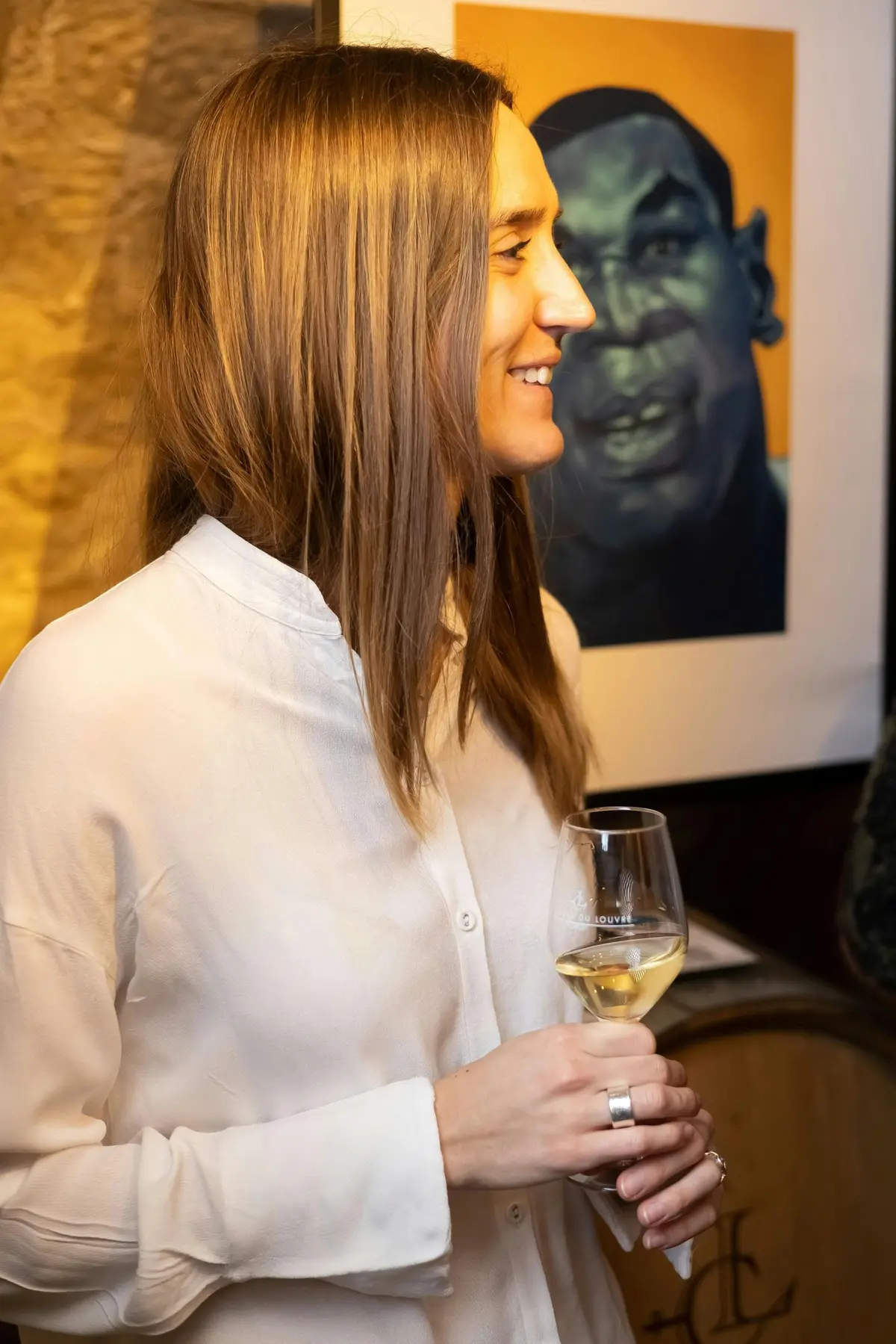 Une personne sourit en tenant un verre, debout près d'un portrait sur un mur. L'arrière-plan présente un éclairage chaud et des murs texturés.