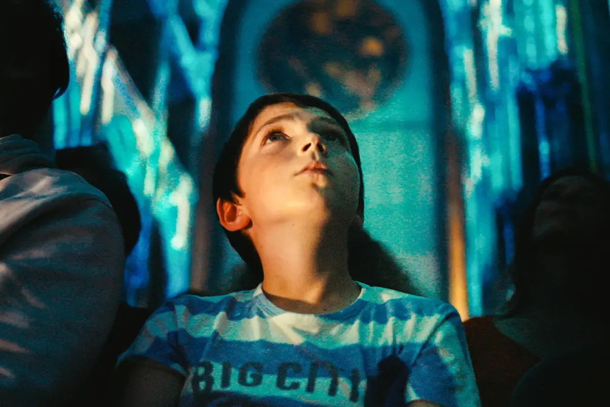 Un jeune lève les yeux dans un décor baigné d'une lumière bleue, avec des motifs complexes en arrière-plan.