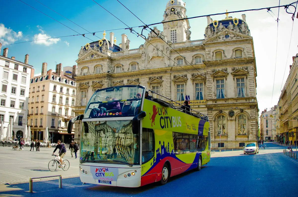 Un bus de la ville de Lyon aux couleurs vives devant un bâtiment historique à l'architecture ornée, par temps clair.
