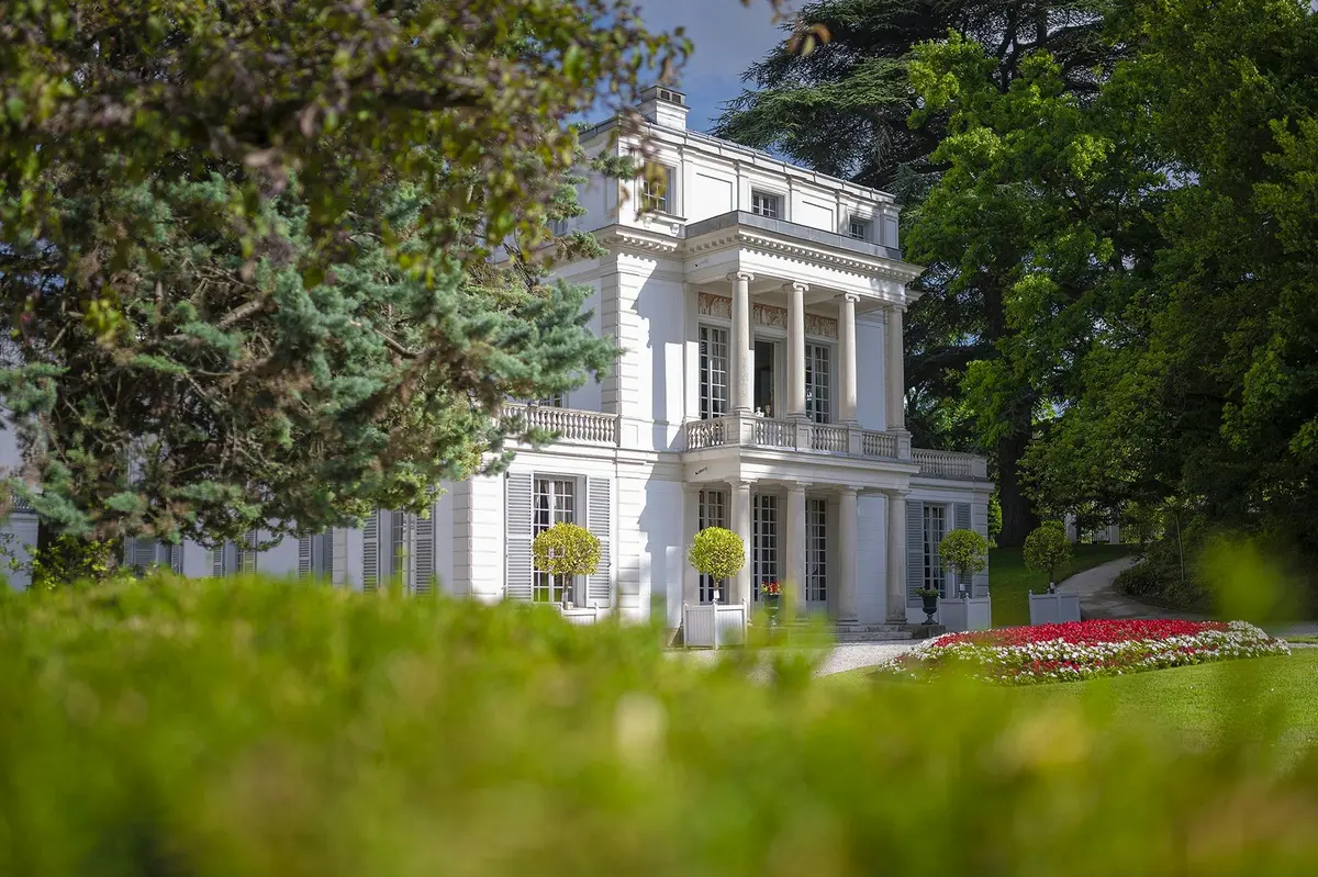 Un bâtiment blanc de style classique avec des colonnes, entouré d'arbres et d'un jardin avec des fleurs et des haies au premier plan.