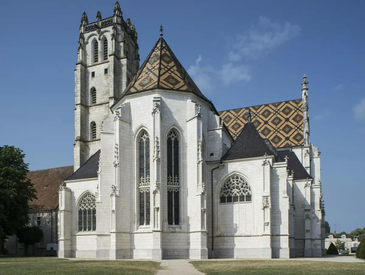 Église de style gothique avec de hautes fenêtres cintrées, des détails ornementaux et un toit à motifs sous un ciel bleu clair.