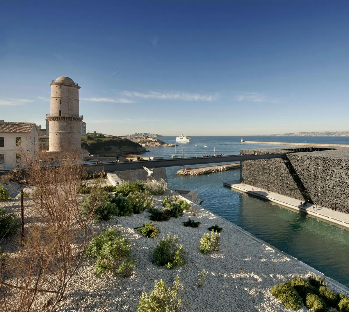 Vieille tour en pierre et b&acirc;timent moderne noir au-dessus de l'eau, avec un rivage rocheux, des plantes et des collines lointaines sous un ciel bleu clair.