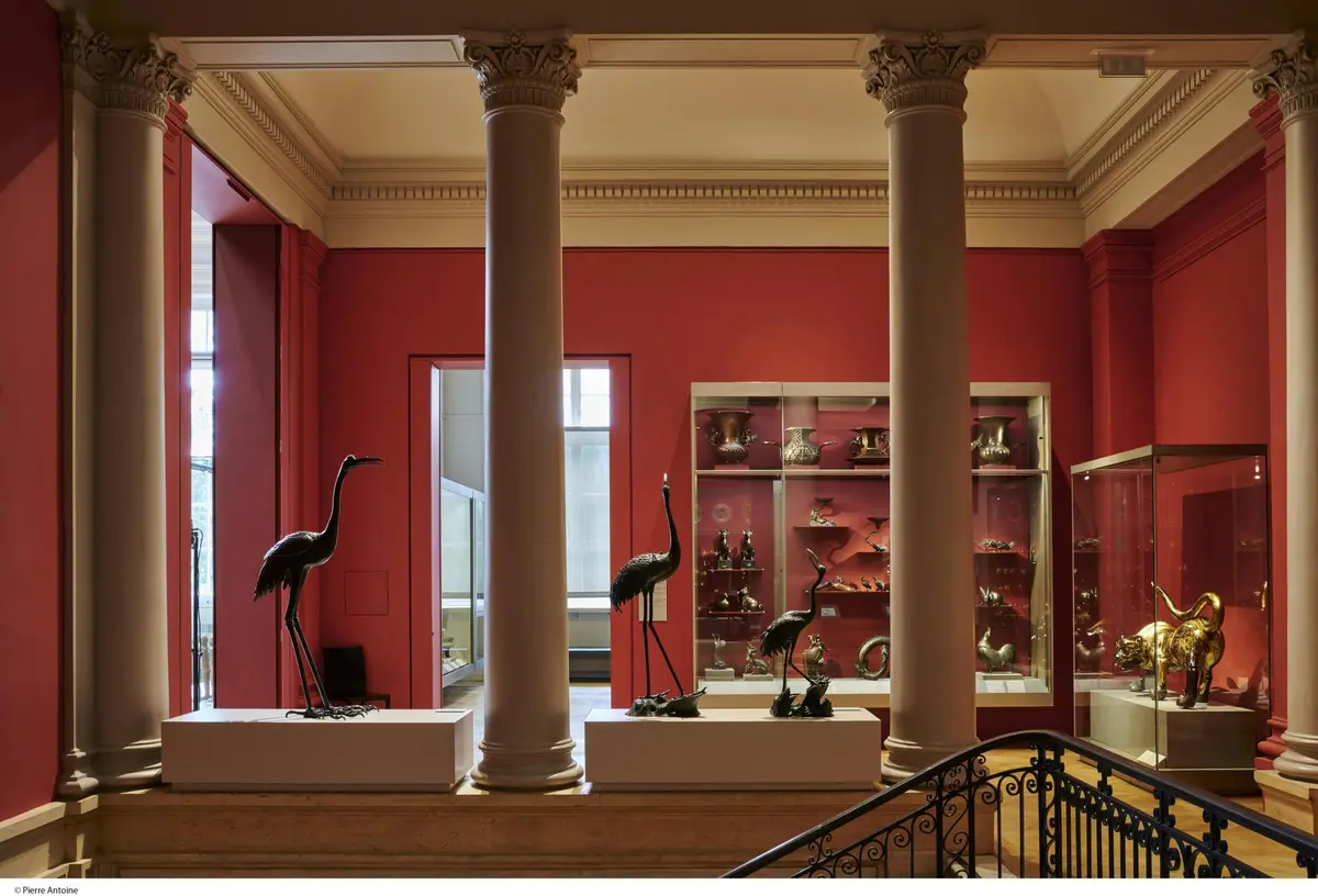 Intérieur du musée avec de hautes colonnes, des murs rouges, des vitrines d'objets en argent et des sculptures d'oiseaux sur des piédestaux.