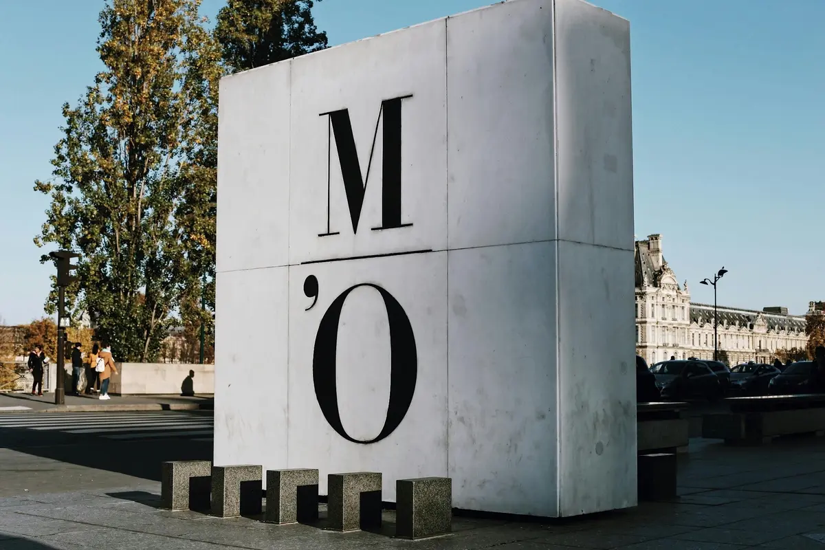 Grande structure cubique blanche portant les lettres M O. Arbres et partie d'un bâtiment en arrière-plan.