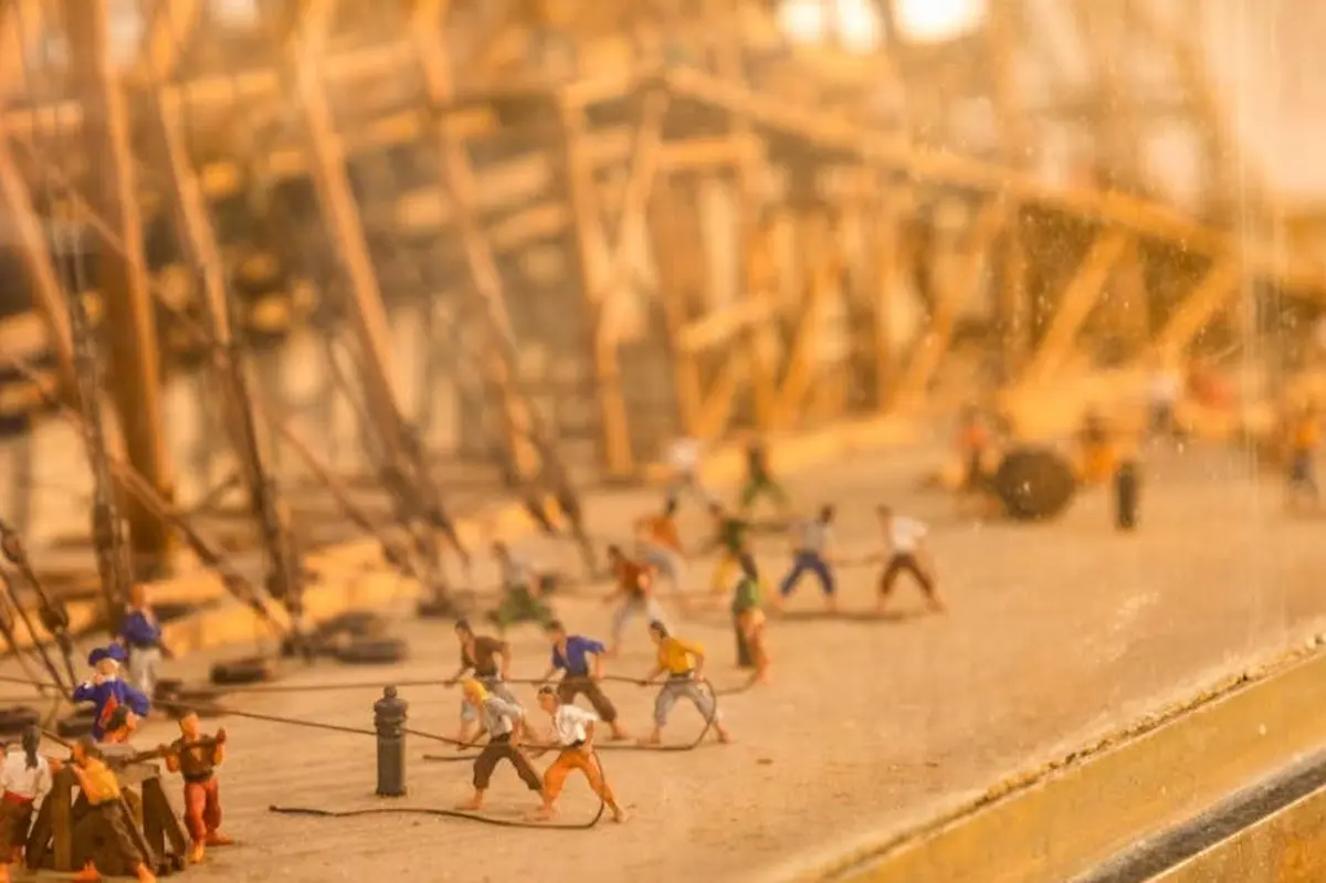 Des figurines représentent des ouvriers tirant des cordes dans un chantier naval en construction, avec des échafaudages en bois à l'arrière-plan.