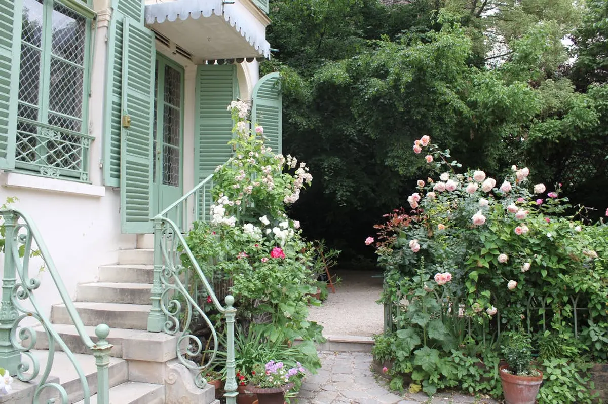 Une charmante entrée de maison aux volets verts, entourée de roses en fleurs et de verdure, menant à une allée de jardin.