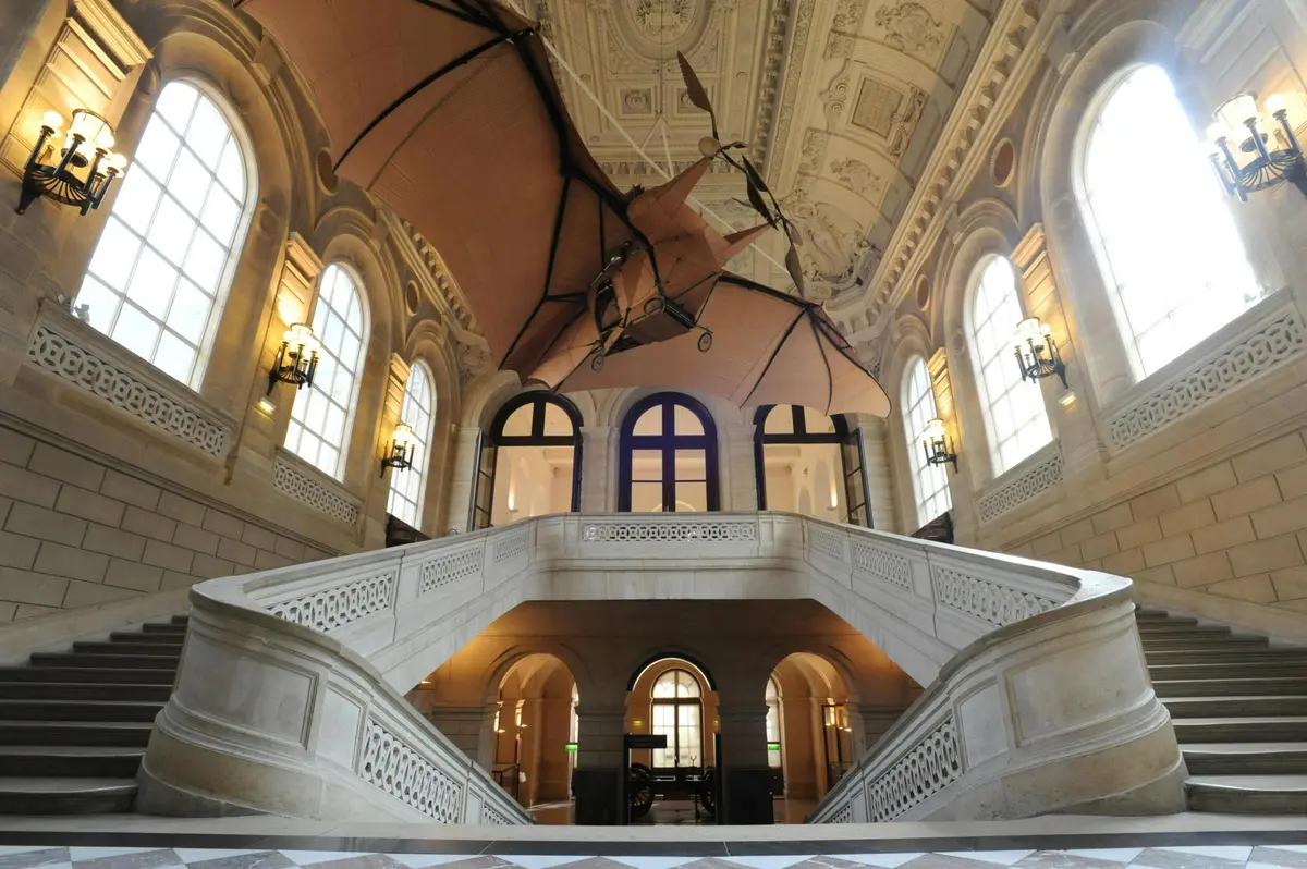 Grand escalier dans un bâtiment historique avec des fenêtres cintrées, un plafond orné et une grande maquette d'avion suspendue.