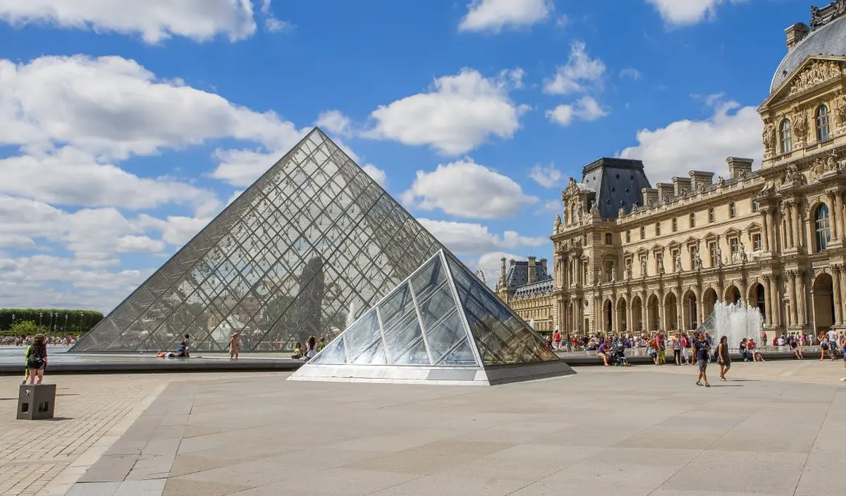 Glass pyramids at the Louvre Museum with people walking and resting around, historic building and blue sky with clouds in the background.