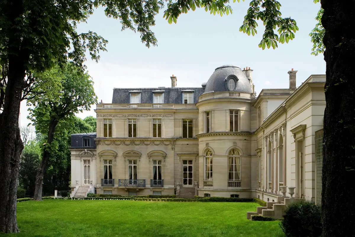 Une grande demeure élégante à l'architecture raffinée, entourée d'arbres et d'une pelouse impeccablement entretenue par temps clair.
