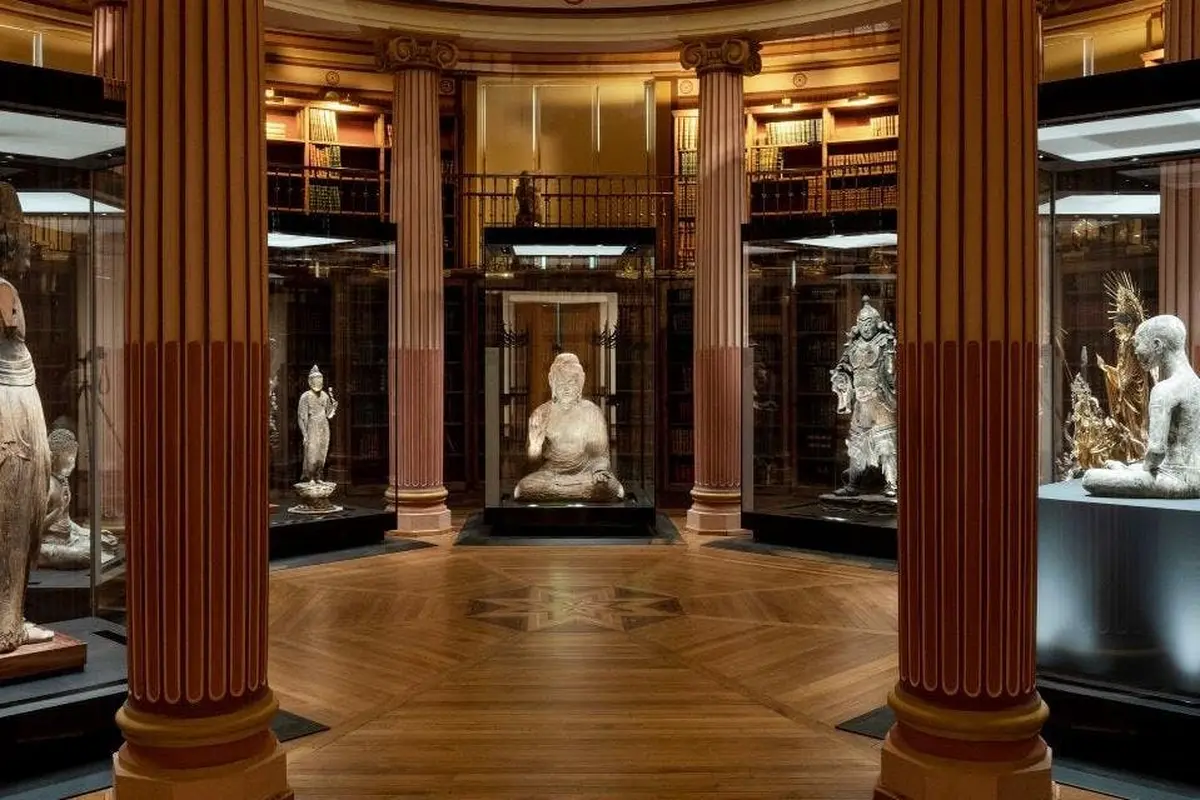 Une salle de musée avec trois sculptures exposées dans des vitrines, entourées de bibliothèques et de colonnes décorées.