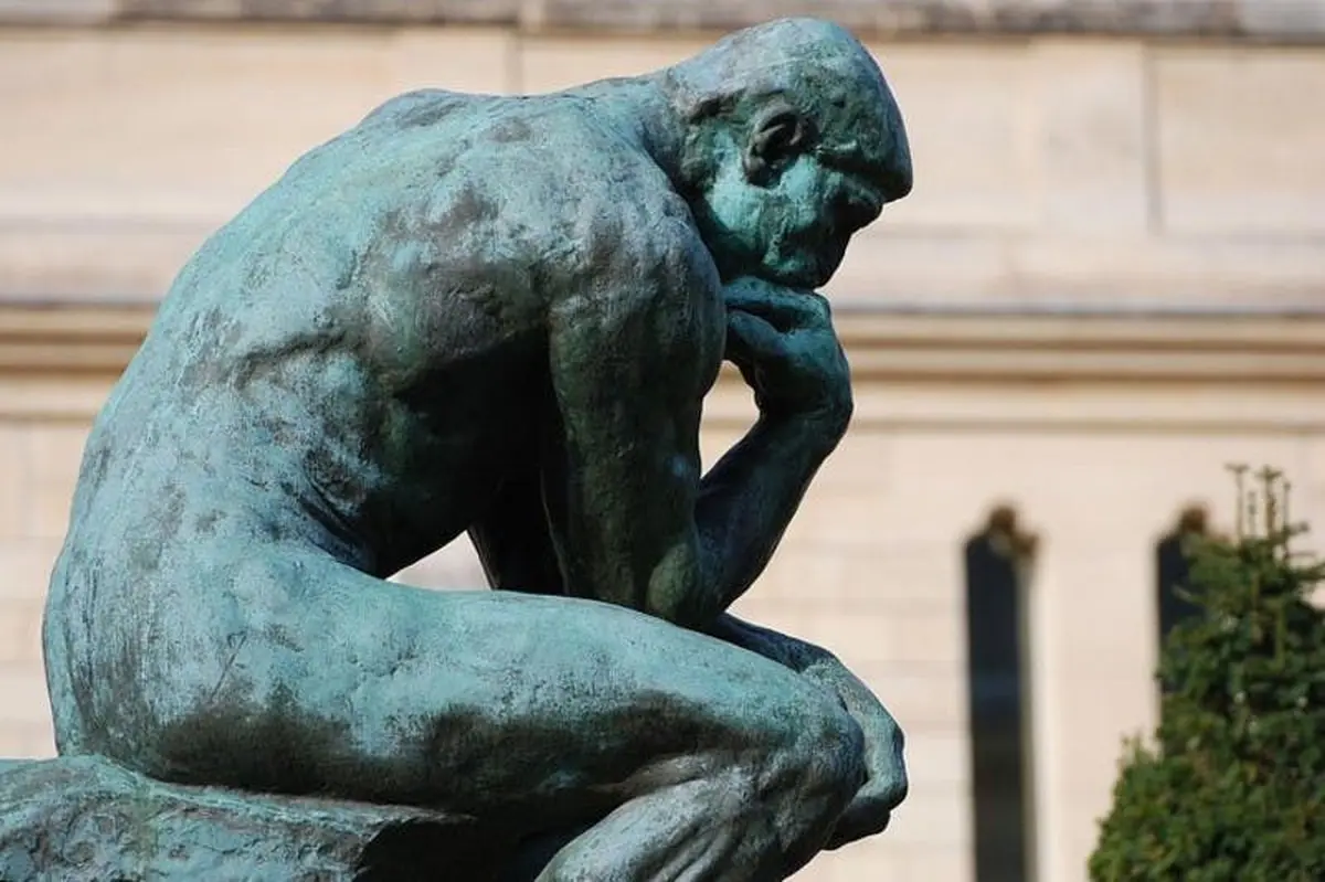 Statue en bronze d'un homme assis, le menton appuyé sur sa main, dans une pose pensive, sur fond d'architecture floue.