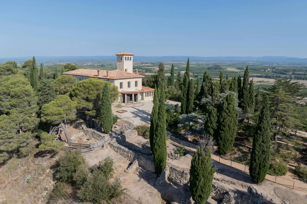 Une villa avec une tour entourée de cyprès et de ruines, dans un paysage de champs et de collines.