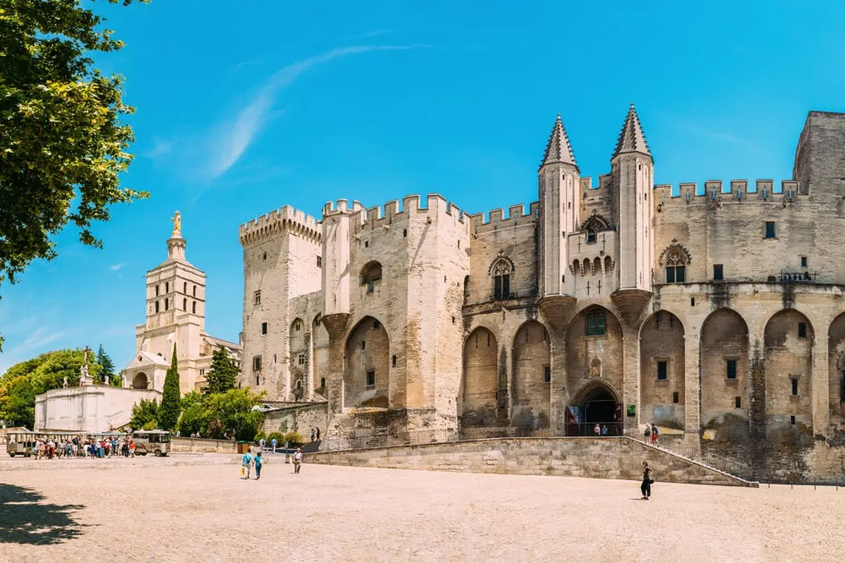Château historique avec de hautes tours et des murs crénelés sous un ciel bleu clair, avec quelques personnes marchant dans la grande cour en contrebas.